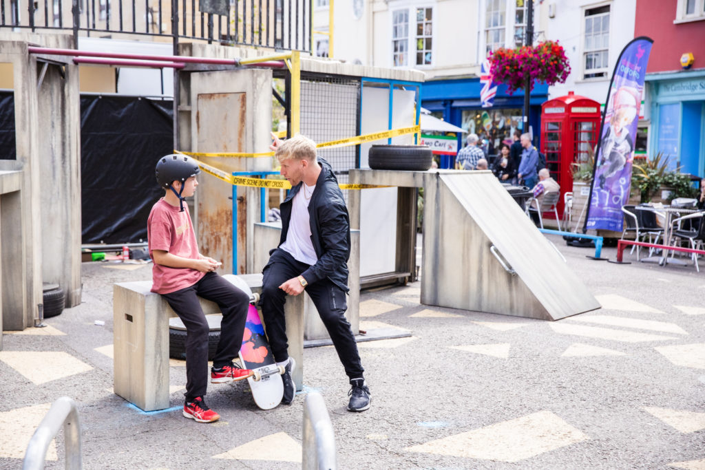 Two seated people, one young with a skateboard wearing a seat pants, t-shirt and a helment, one old wearing sweat pants t- shirt and bomber jacket