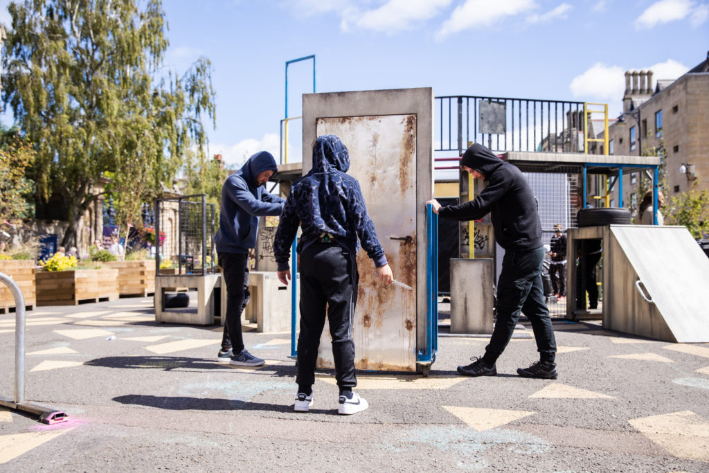 Two men in hoodies and sweatpants hold a door in position. in front of the door a young person in sweatpants and a hoodie, faces away from the camera, towards the door, knife in hand.