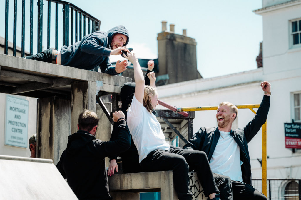 Five people, two sitting on a platform one with arms raised in celebration, one lying on a ledge above, and two on either side of the platform. All are wearing sweat pants and t-shirts, some are wearing hoodies.